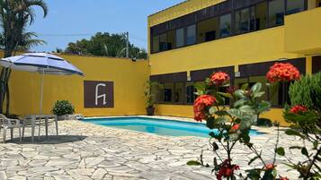 Zona de piscina con baldosas de piedra, un edificio amarillo con letrero 'H' y flores rojas en primer plano.
