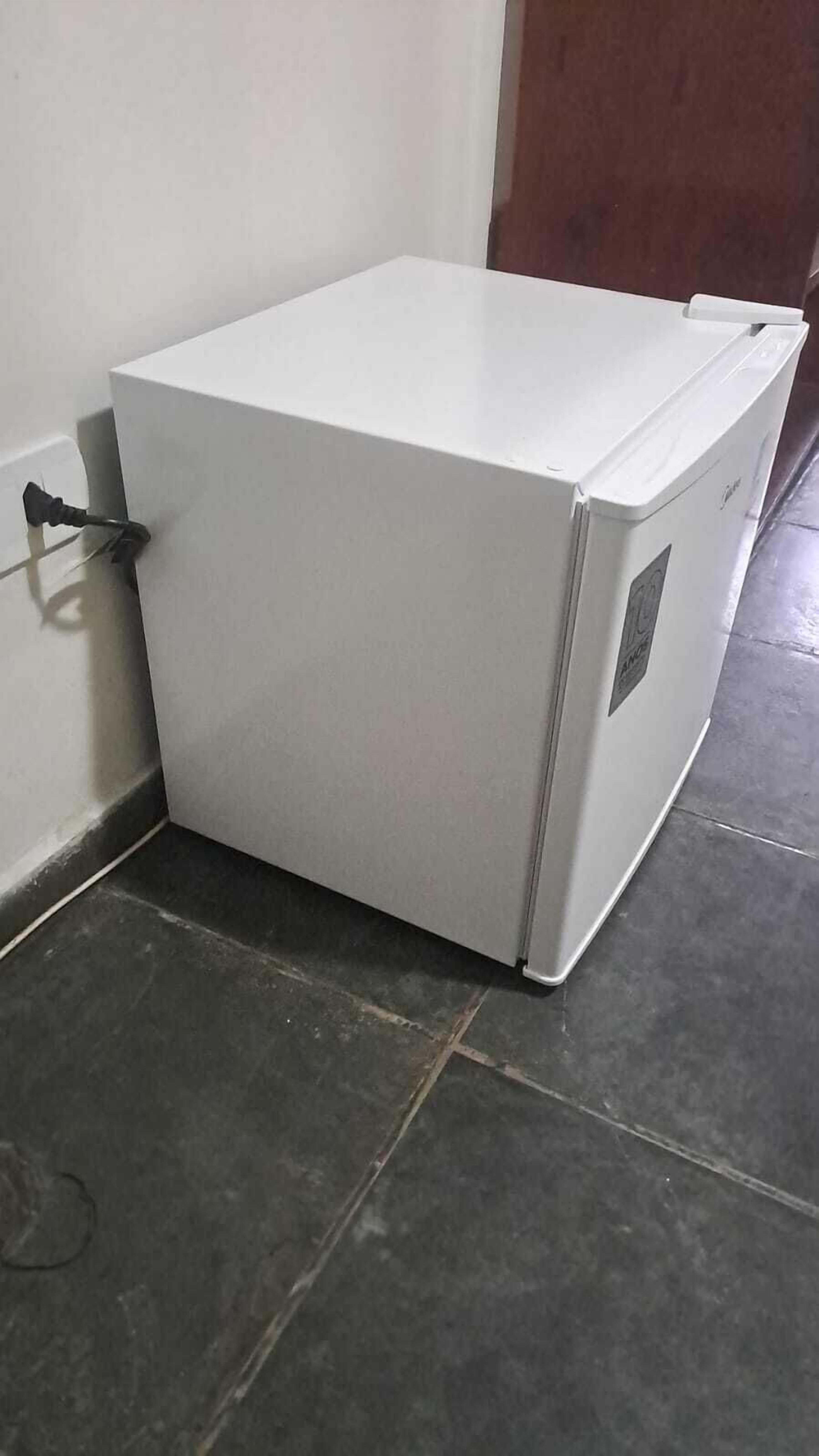 A white refrigerator in a corner of a room with dark tiled flooring.