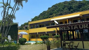 A two-story yellow building with a sign reading "RESTAURANT" on the ground floor. There is a pool area with lounge chairs and umbrellas. - Pousada Cantinho do Mar  (Guarujá)