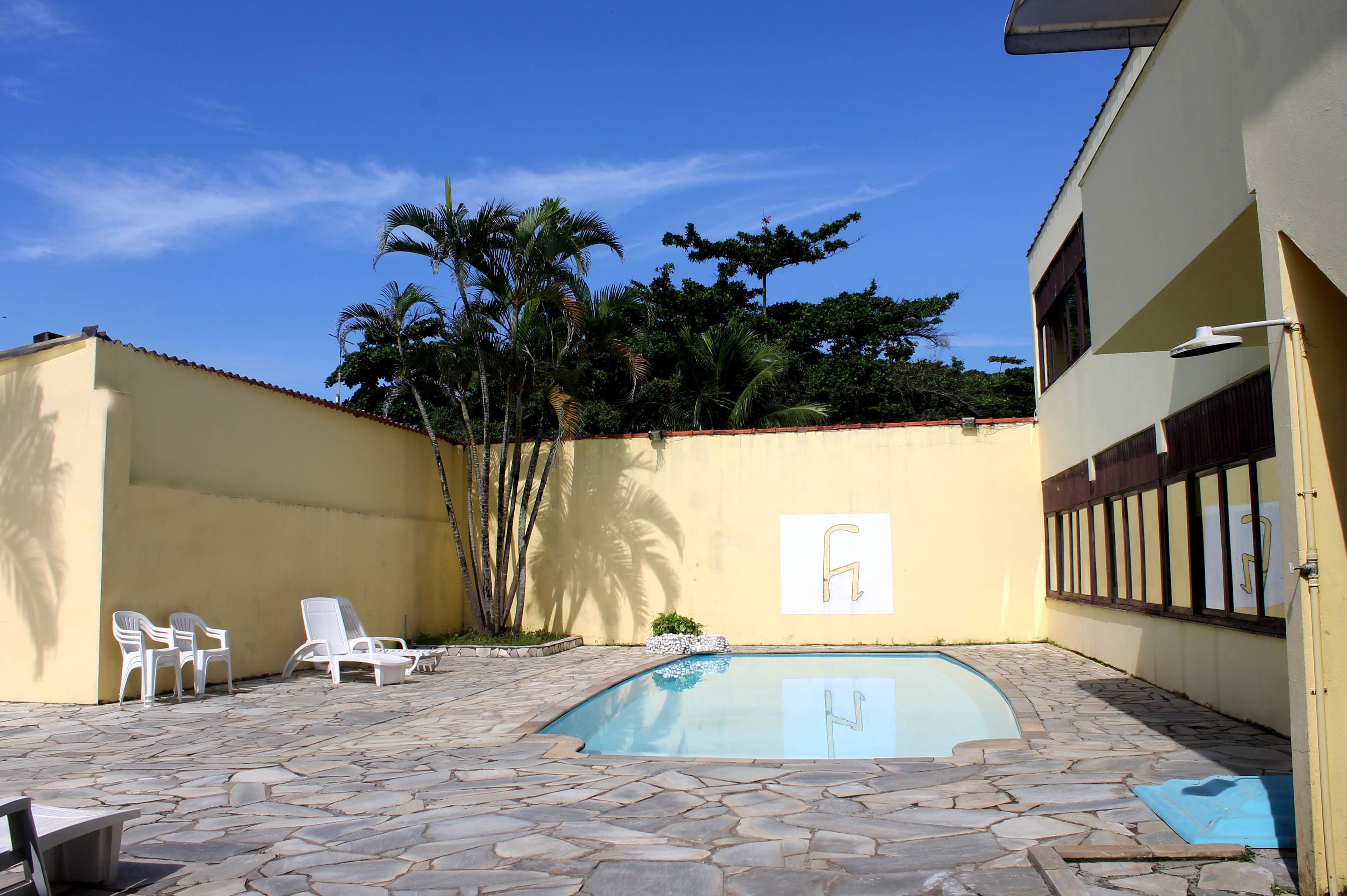 Una piscina con baldosas de piedra, sillas de descanso blancas y un edificio amarillo con ventana y puerta.