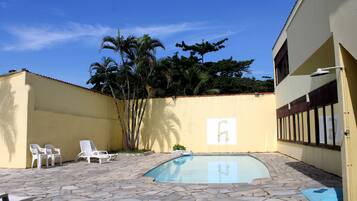 Una piscina con baldosas de piedra, sillas de descanso blancas y un edificio amarillo con ventana y puerta.