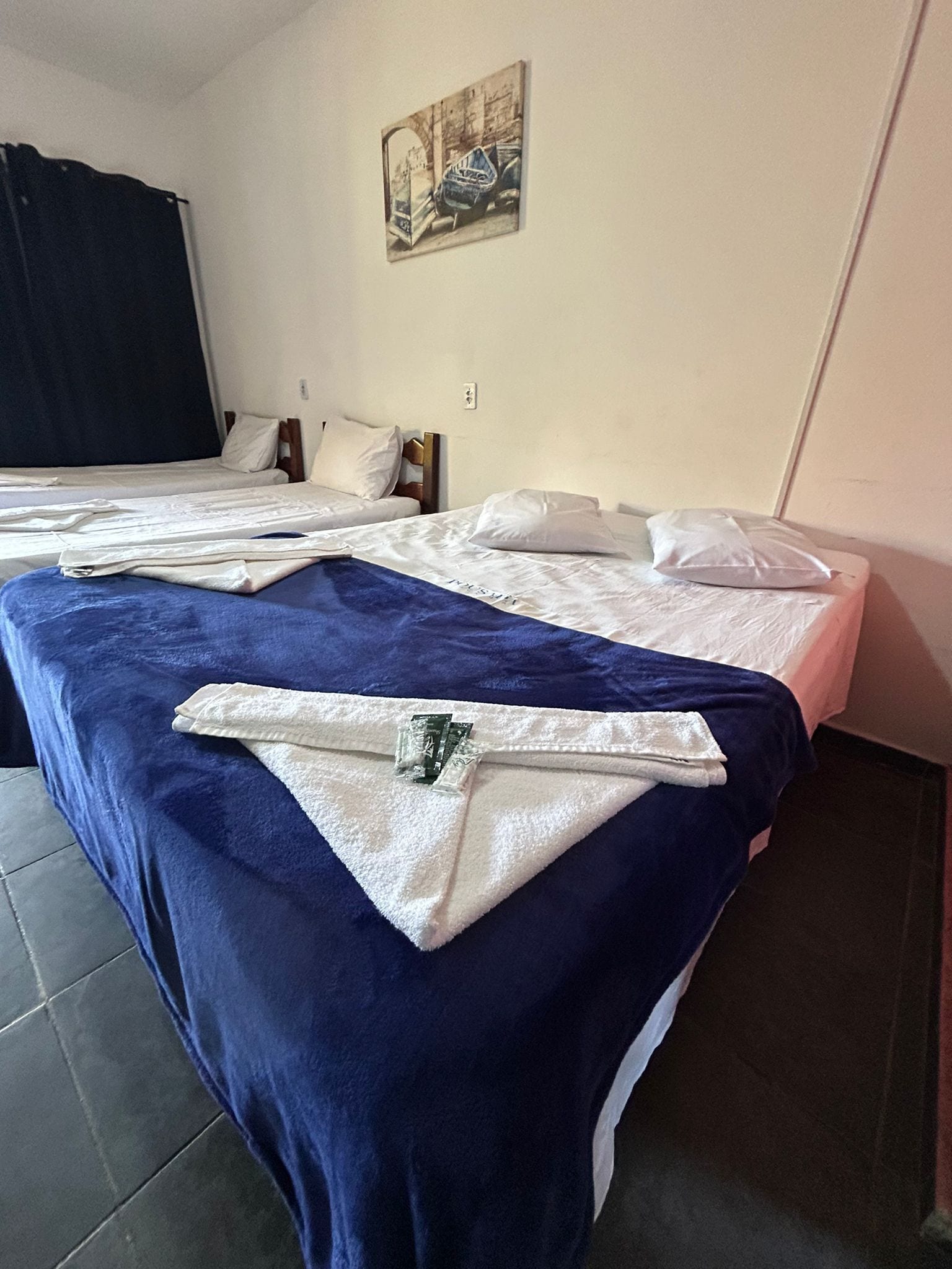 A hotel room with two beds, each with white linens and pillows, a blue blanket on the left bed, and a painting on the wall.