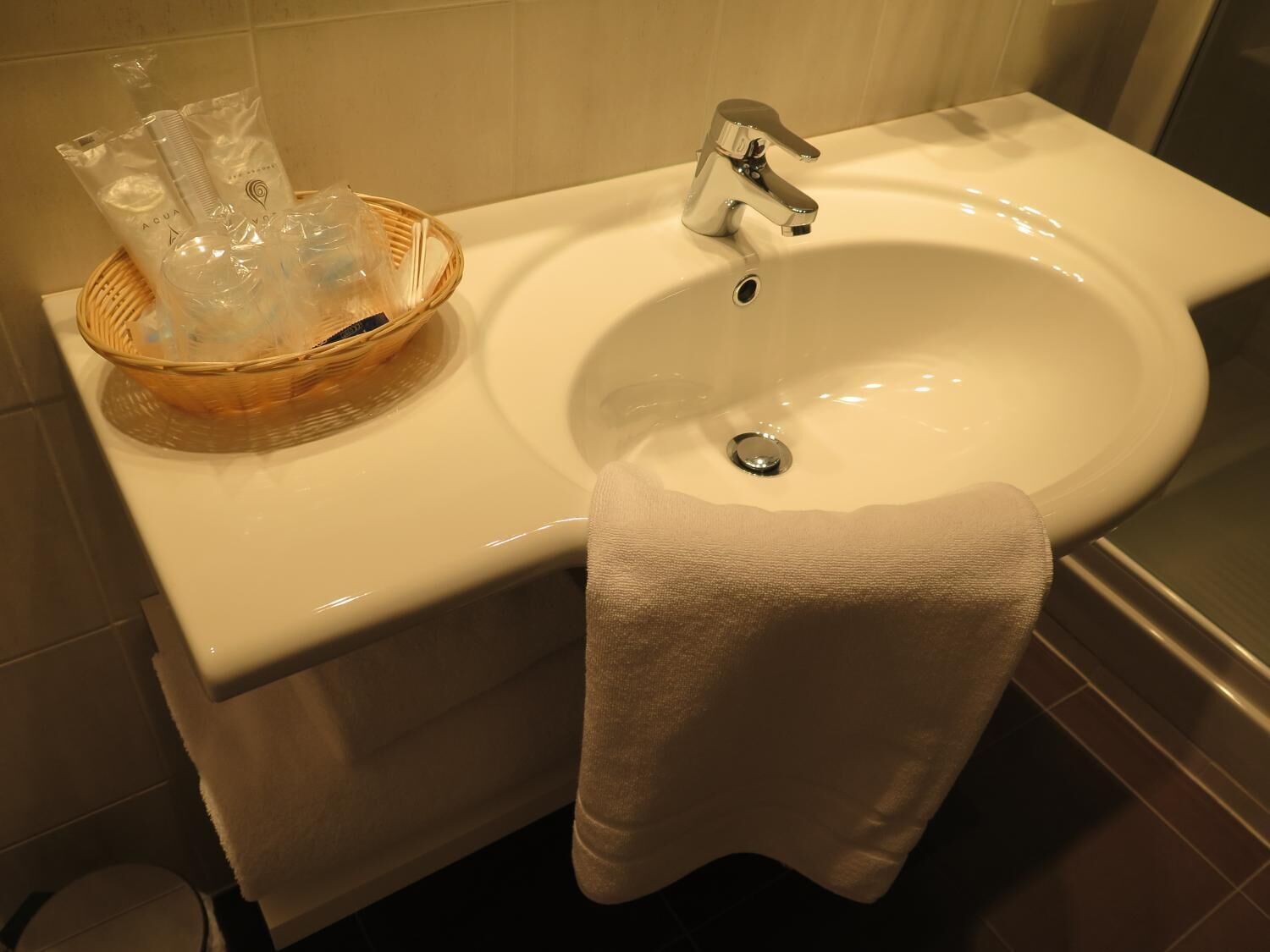 Un baño con un lavabo blanco, un grifo cromado, una toalla blanca colgada en la pared y una cesta de mimbre con artículos de aseo.