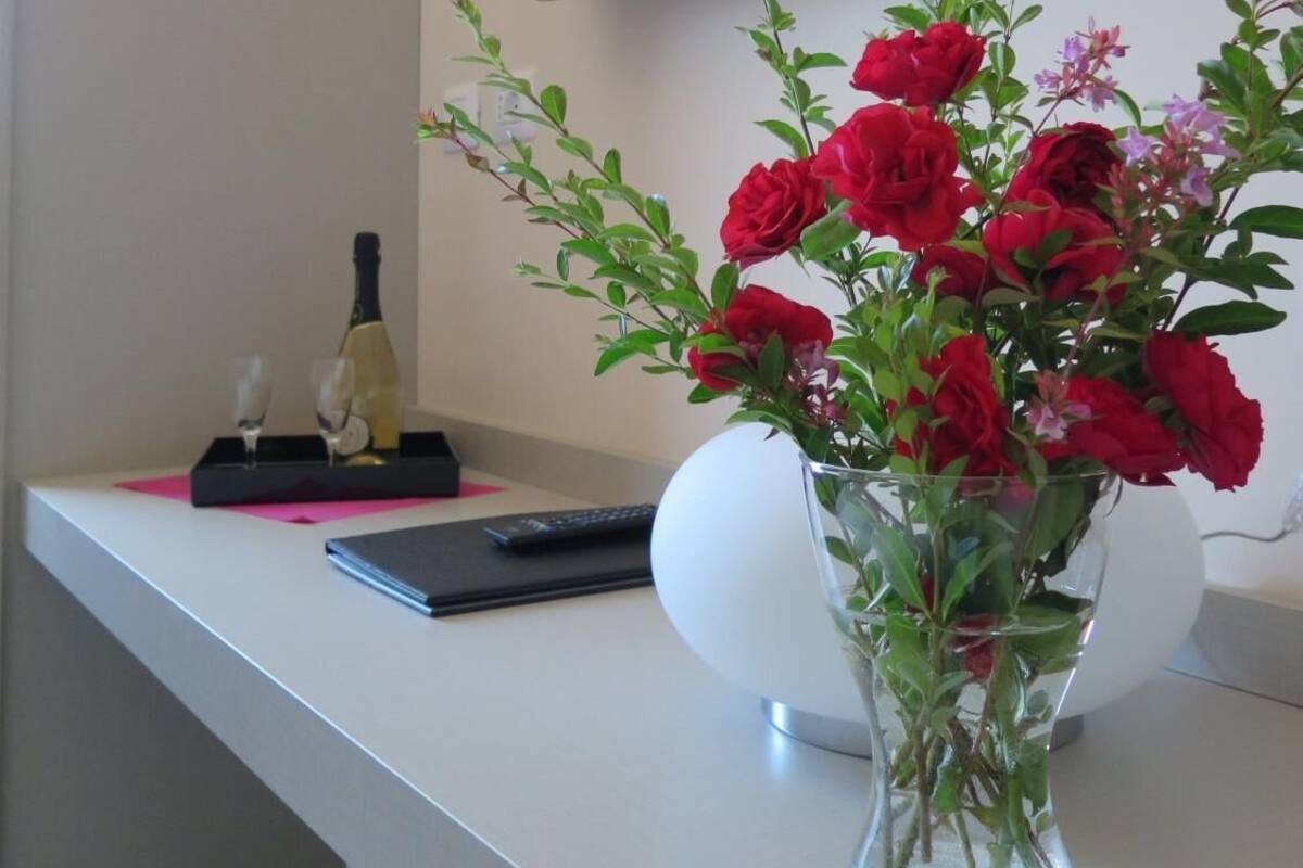A modern room with a flat-screen TV mounted on the wall, a white desk with a vase of red flowers, and a bottle of champagne.