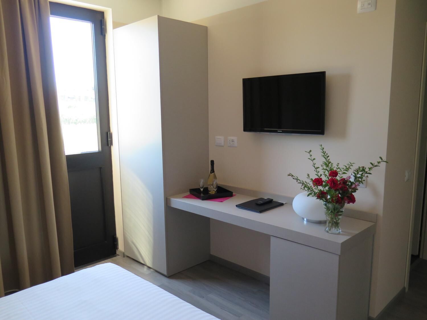Habitación de hotel con cama, escritorio con televisor de pantalla plana, florero con flores y ventana con cortinas.