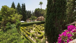 Un jardín ajardinado con setos cuidados, diversas especies de árboles y un edificio con tejado de tejas.