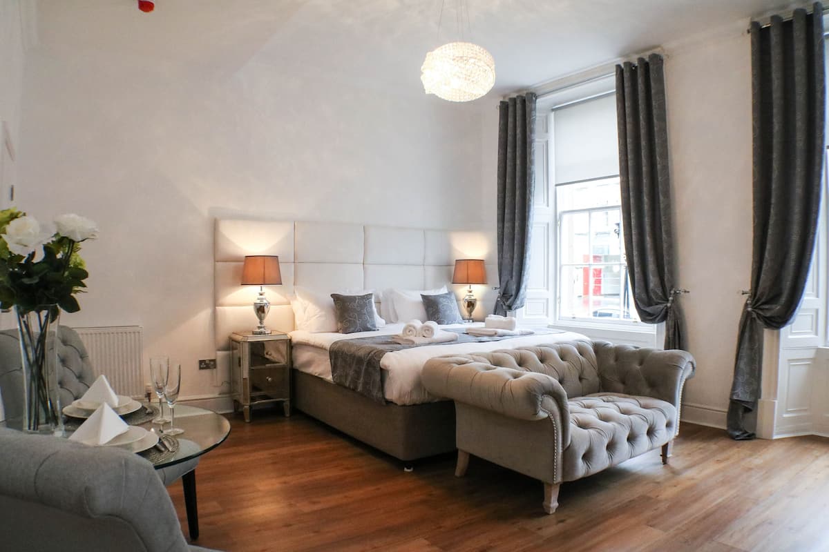 A bedroom with a large bed, bedside lamps, a tufted armchair, a round table with a vase and napkins, and a window with curtains.