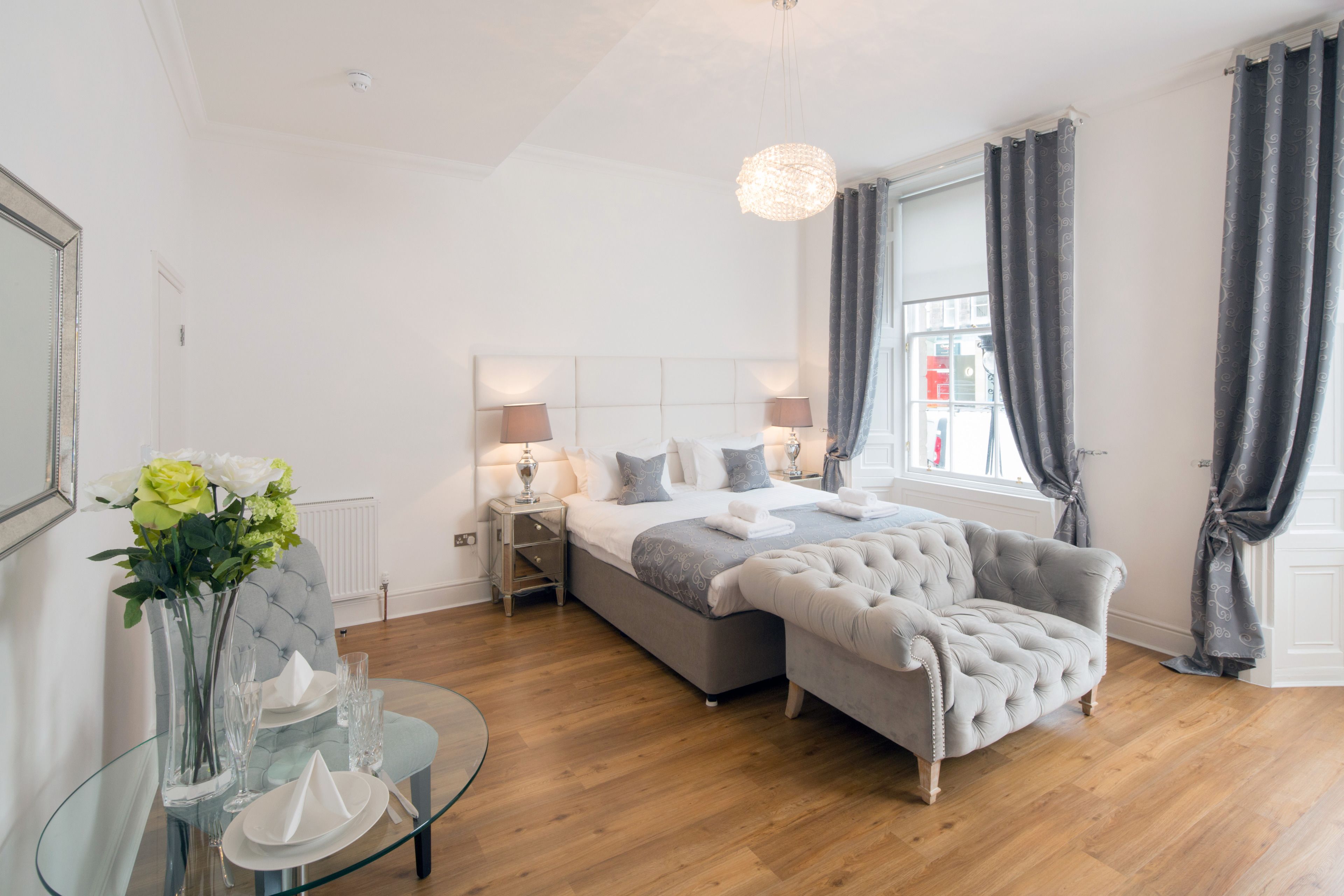 A modern bedroom with a large bed, a tufted armchair, a glass table with a vase of flowers, and a view of the city through the window.