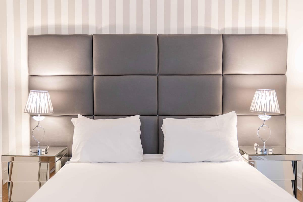 A neatly made bed with a large padded headboard, two bedside lamps, and a glass side table.