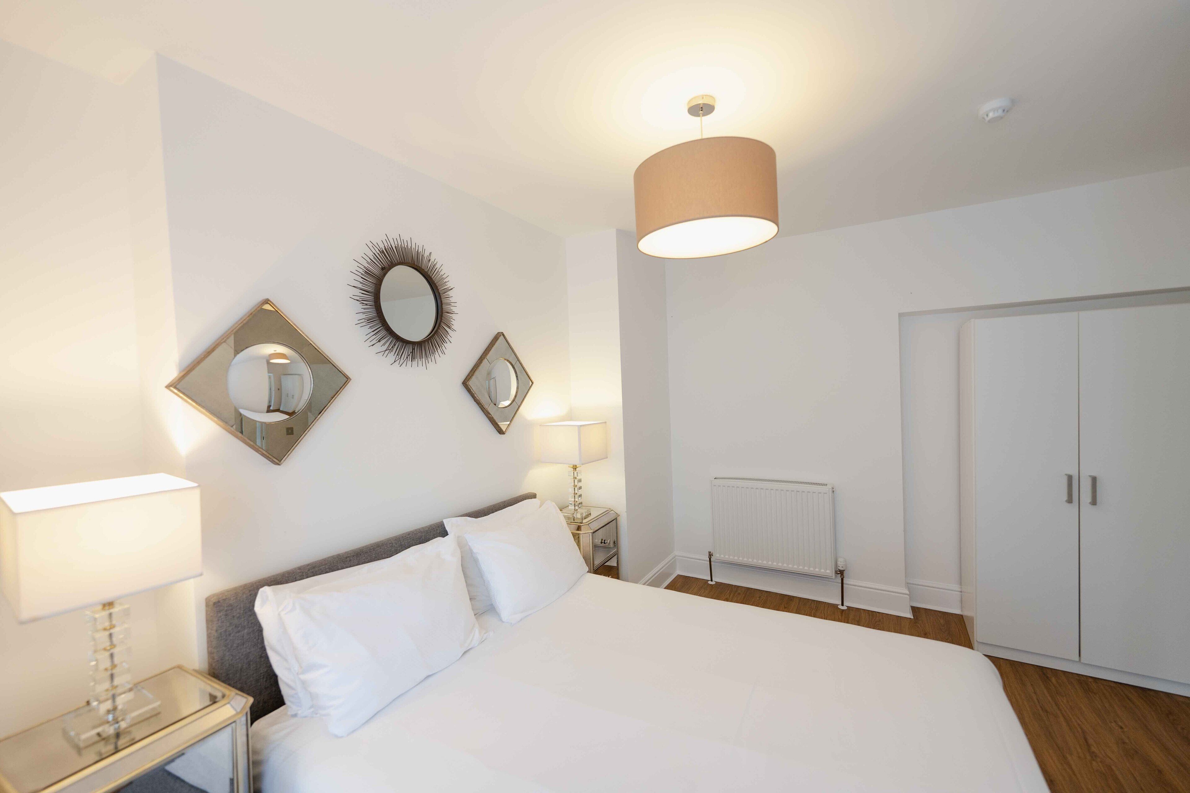 A hotel room with a bed, bedside lamps, a mirror, a radiator, and a wardrobe.