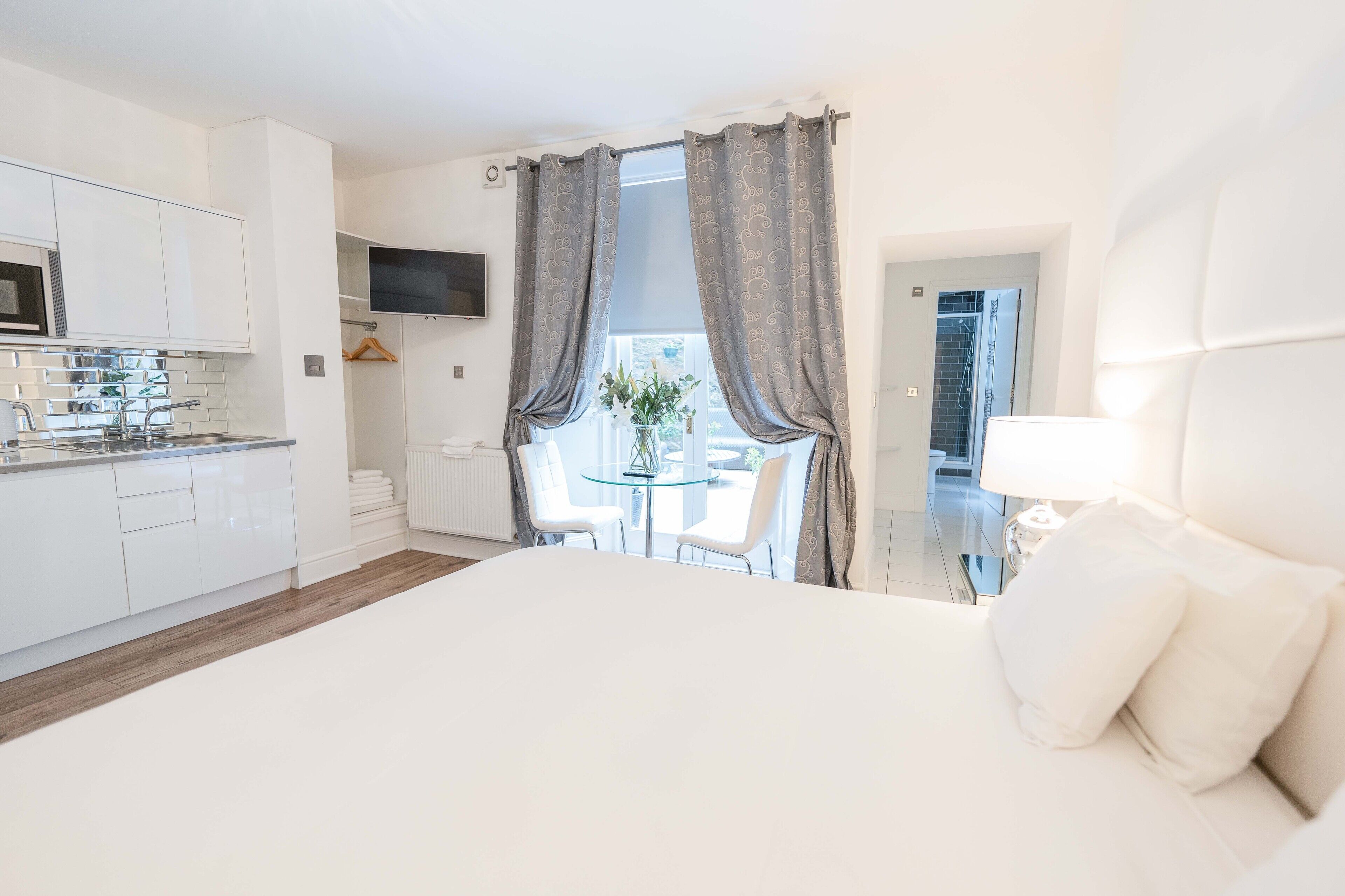 A modern hotel room with a large bed, a glass table with chairs, a kitchen area, and a TV mounted on the wall.