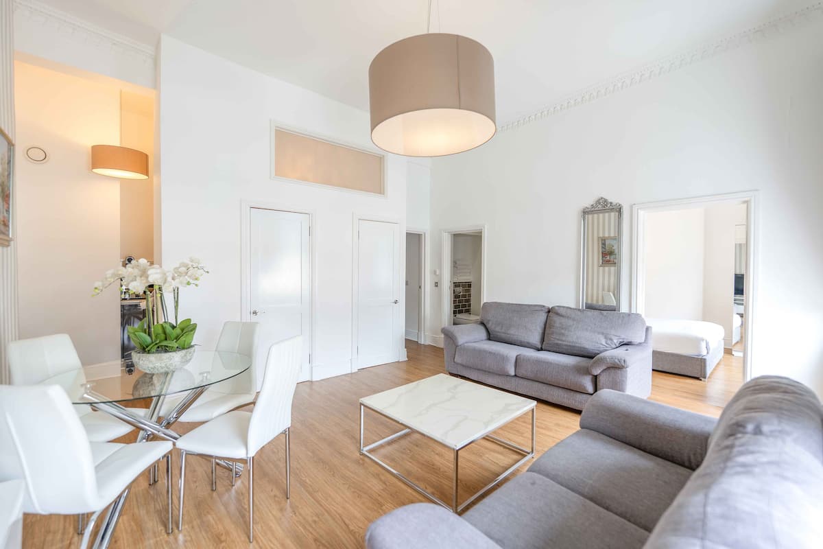 A modern living room with a glass dining table, grey sofas, and a large hanging light fixture.