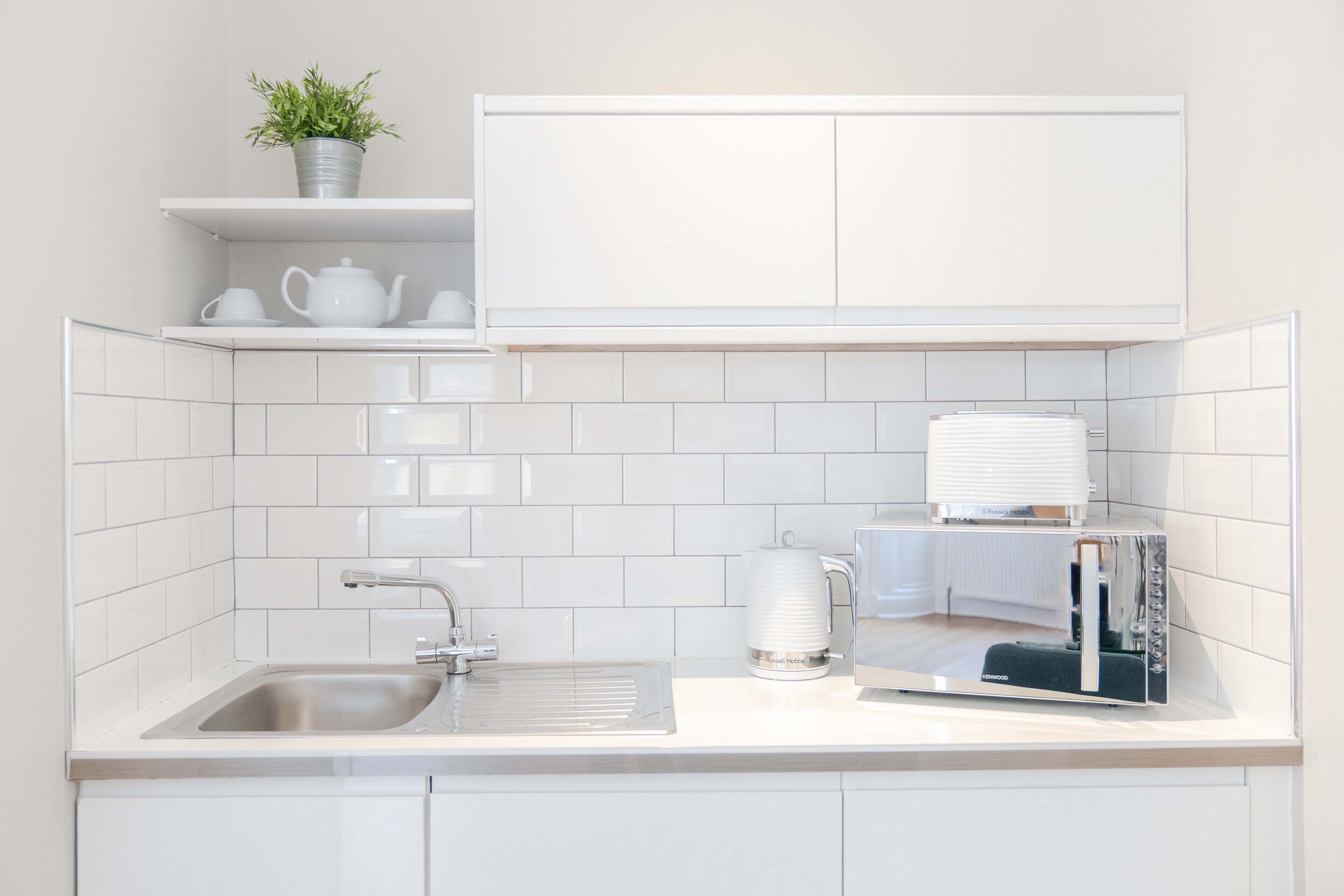 Una cocina moderna con muebles blancos, un lavamanos de acero inoxidable, una tetera y un microondas.