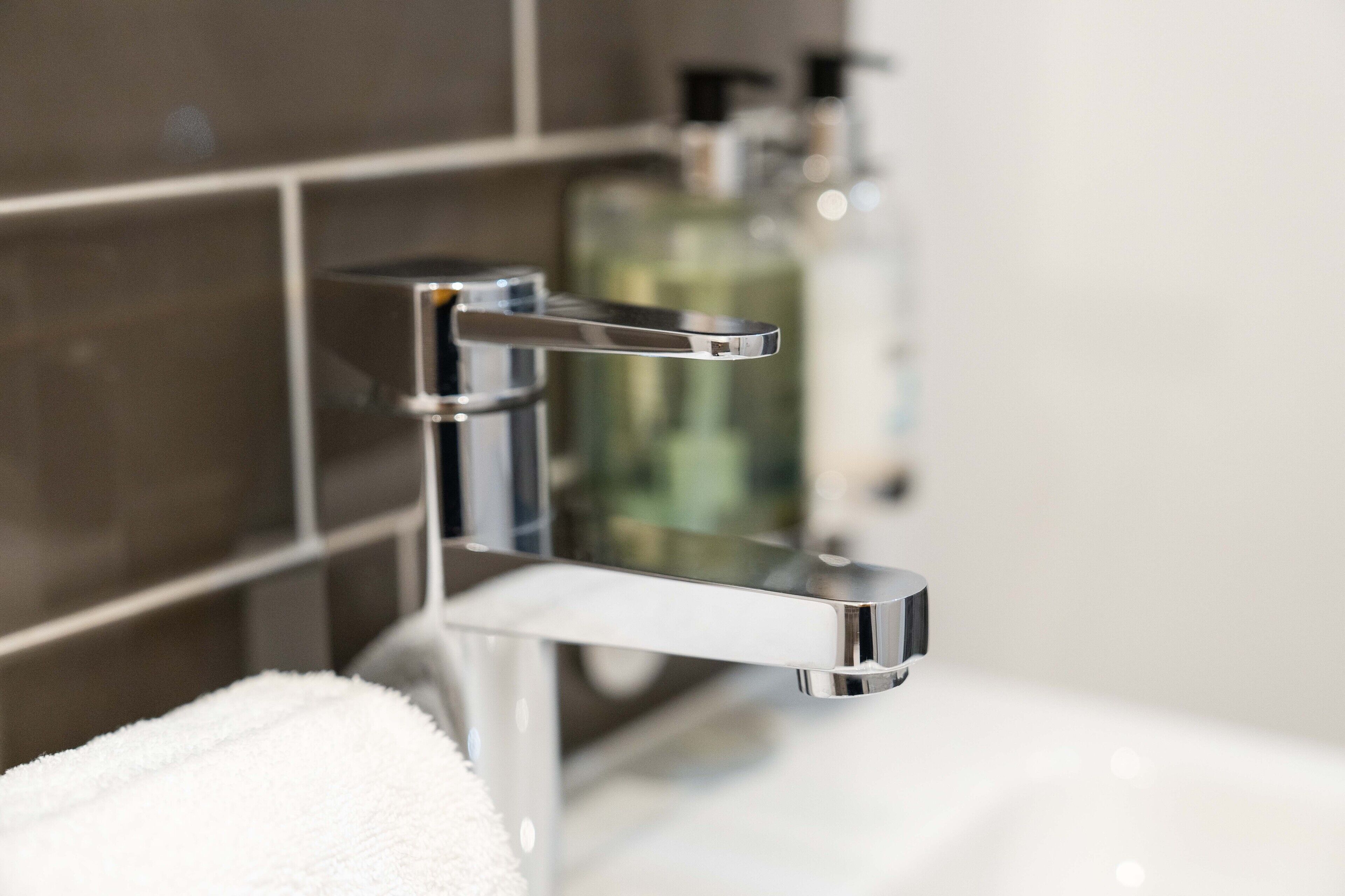 Een moderne badkamer met een chromen kraan, een witte wastafel en een glazen fles vloeibare zeep op de achtergrond.