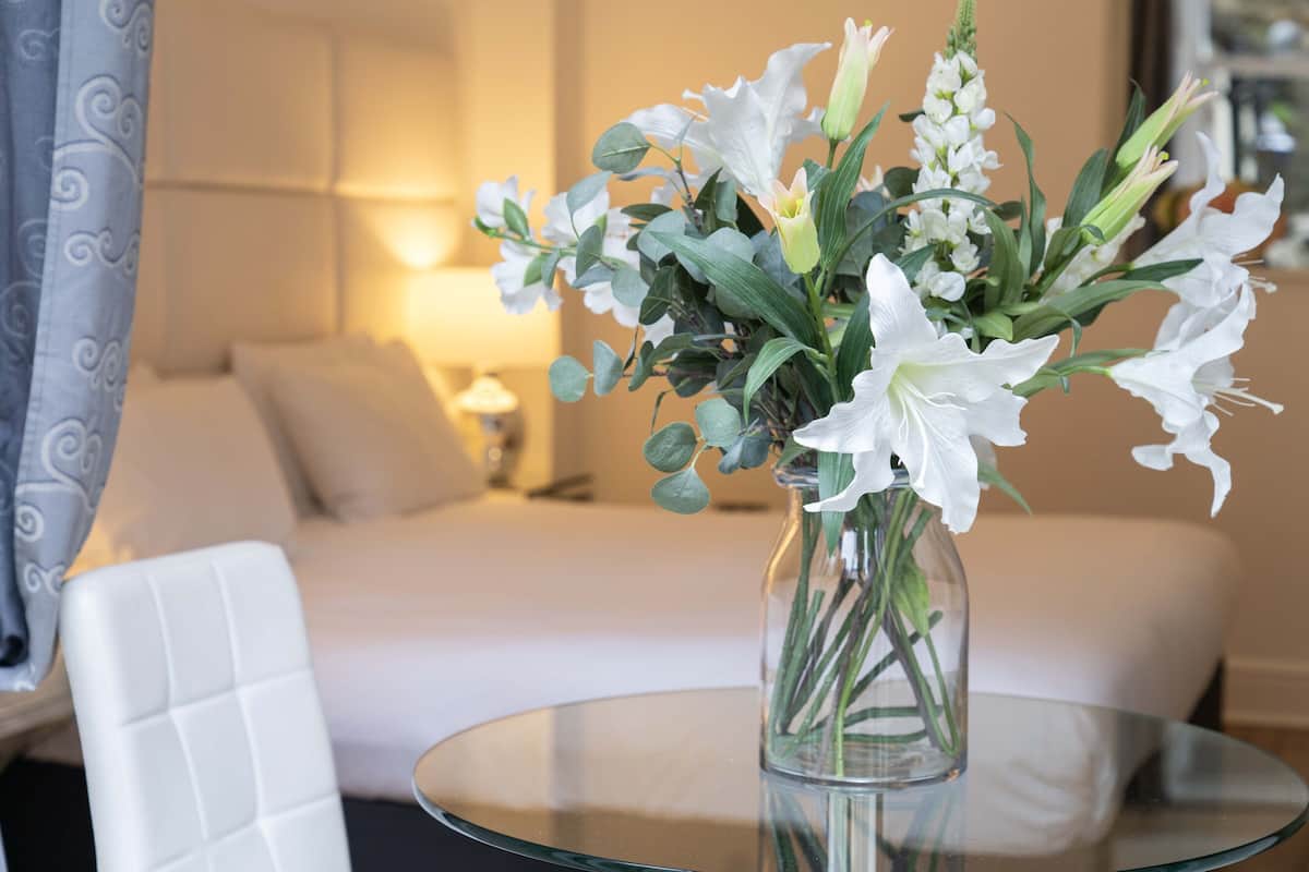 A hotel room with a bed, a glass table with a vase of flowers, and a white chair.