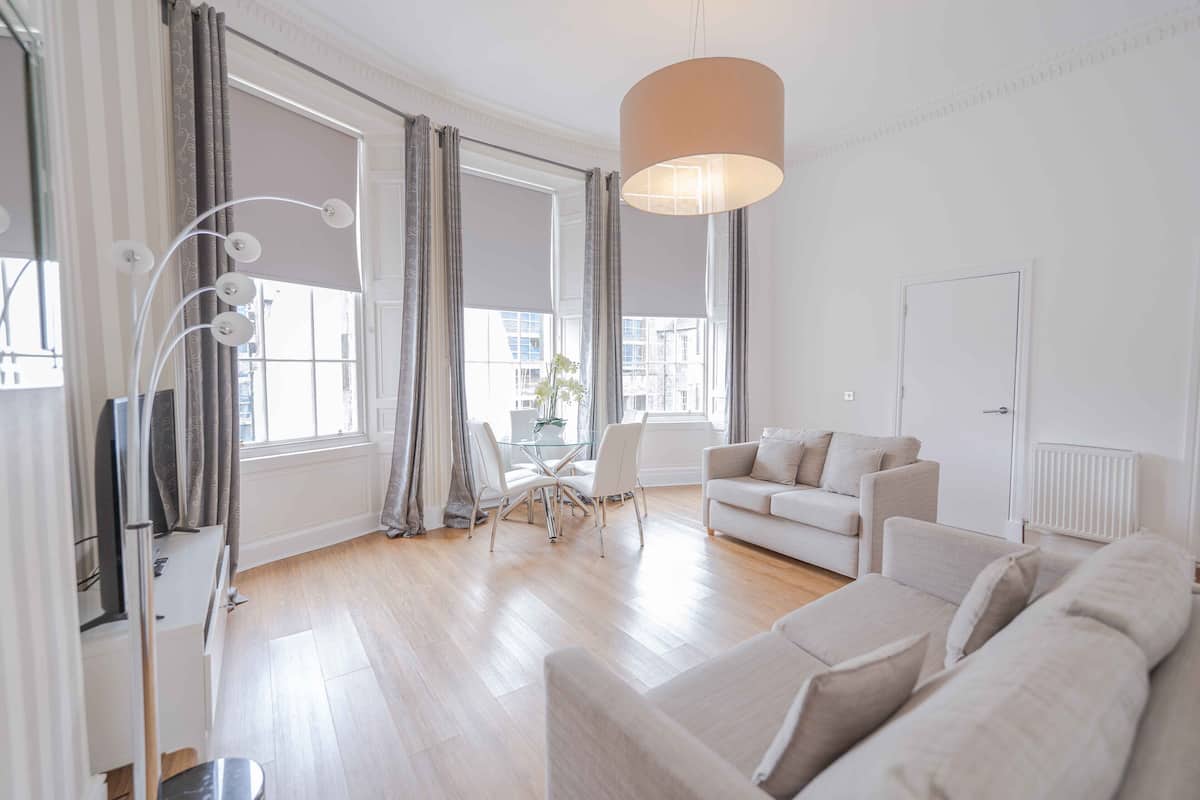 A modern living room with a sofa, a dining table with chairs, and a large window with curtains.
