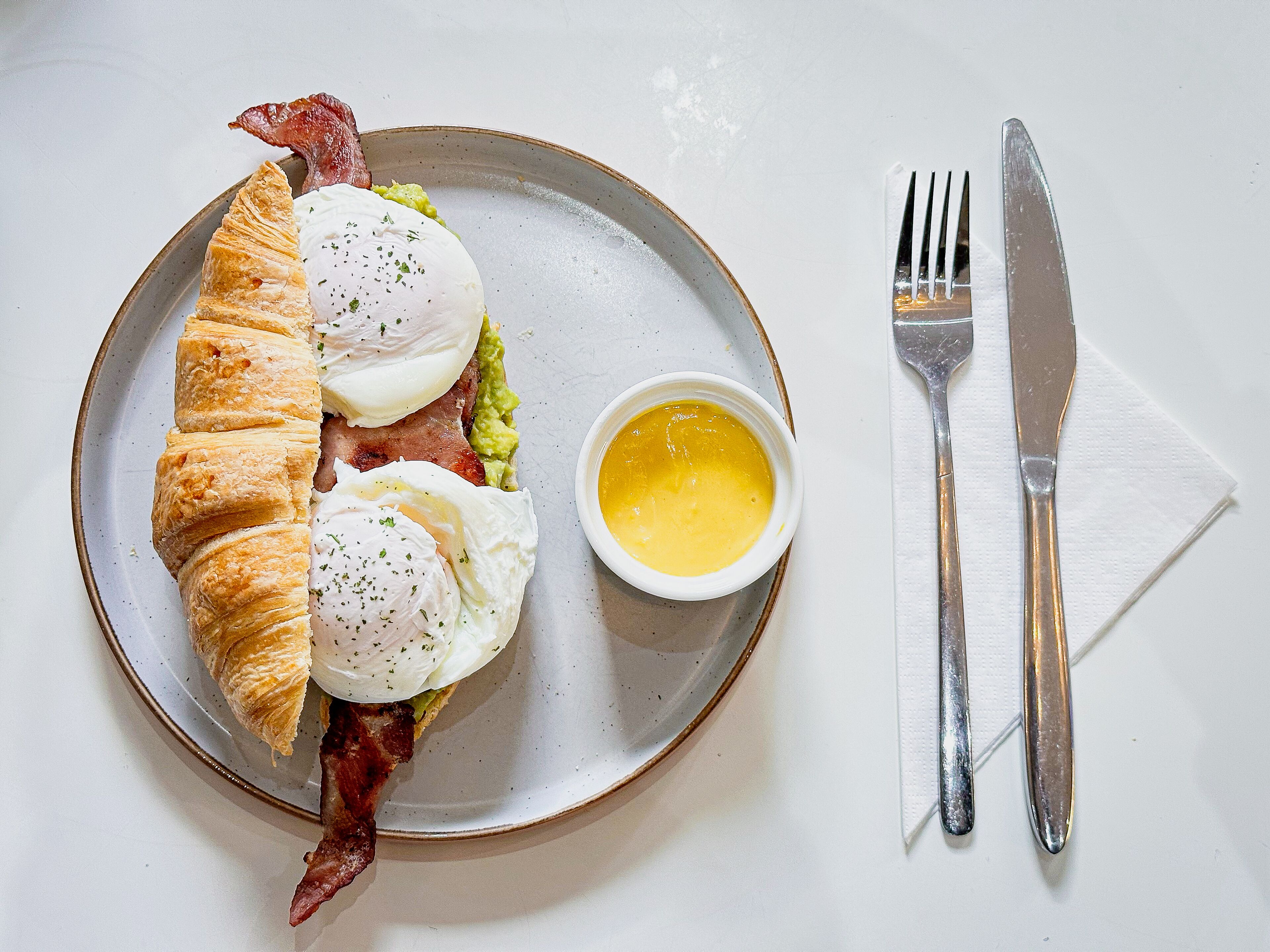 Un plato con un croissant, dos huevos pochados y tocino, acompañado de un recipiente pequeño de salsa, sobre una mesa blanca con servilleta y cubiertos.