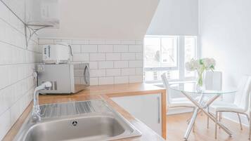 Una cocina moderna con azulejos blancos, un lavamanos de acero inoxidable, un tostador empotrado y una mesa de vidrio con sillas blancas.