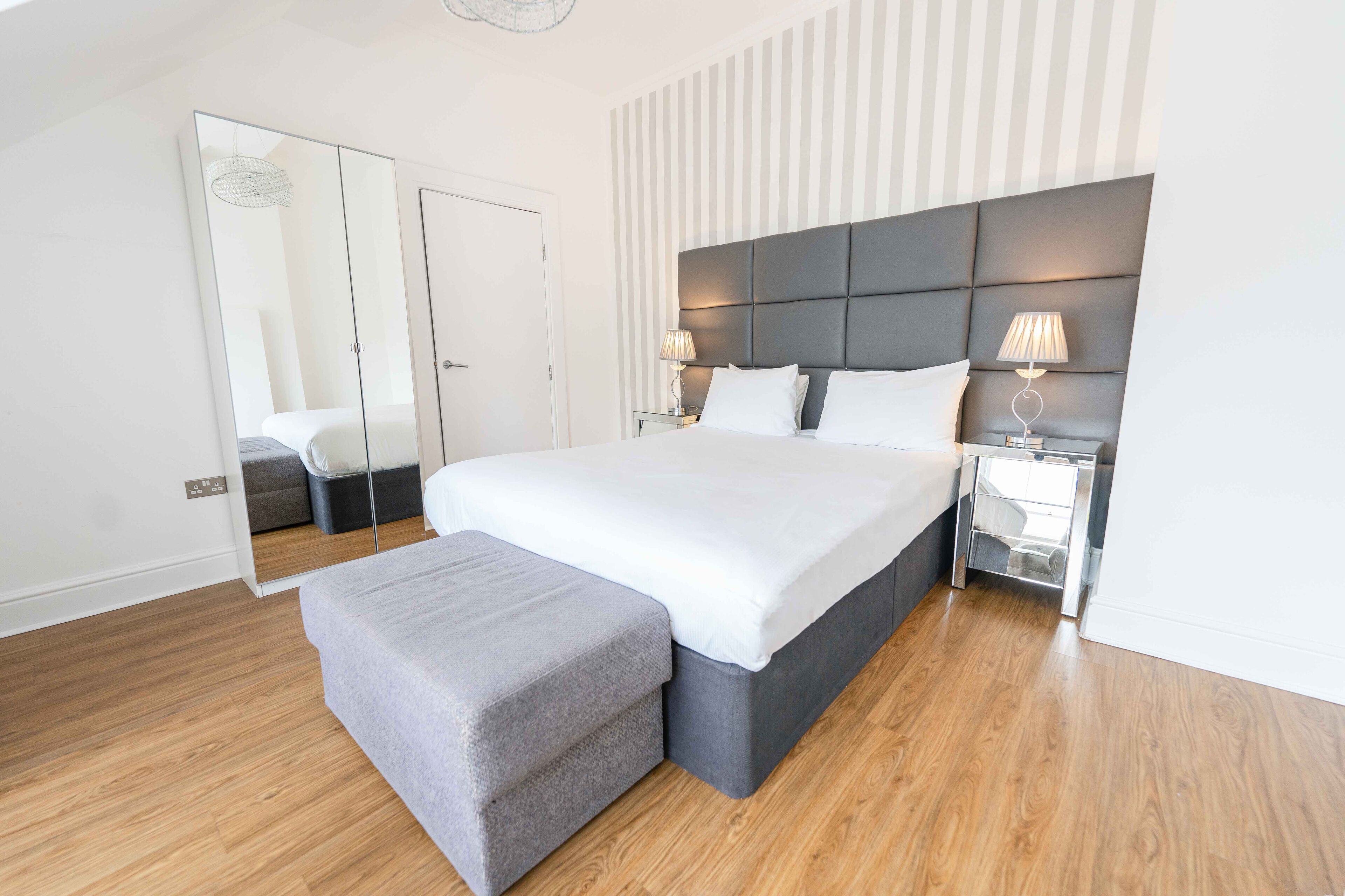A modern bedroom with a large bed, bedside tables, a bench, a mirror, and a striped wall.