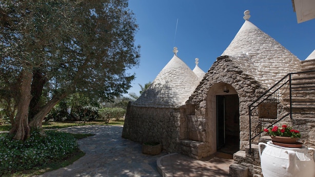 A pathway leading to stone buildings with conical roofs, surrounded by greenery and a large white urn with flowers. - Villa Ricci (Ceglie Messapica)