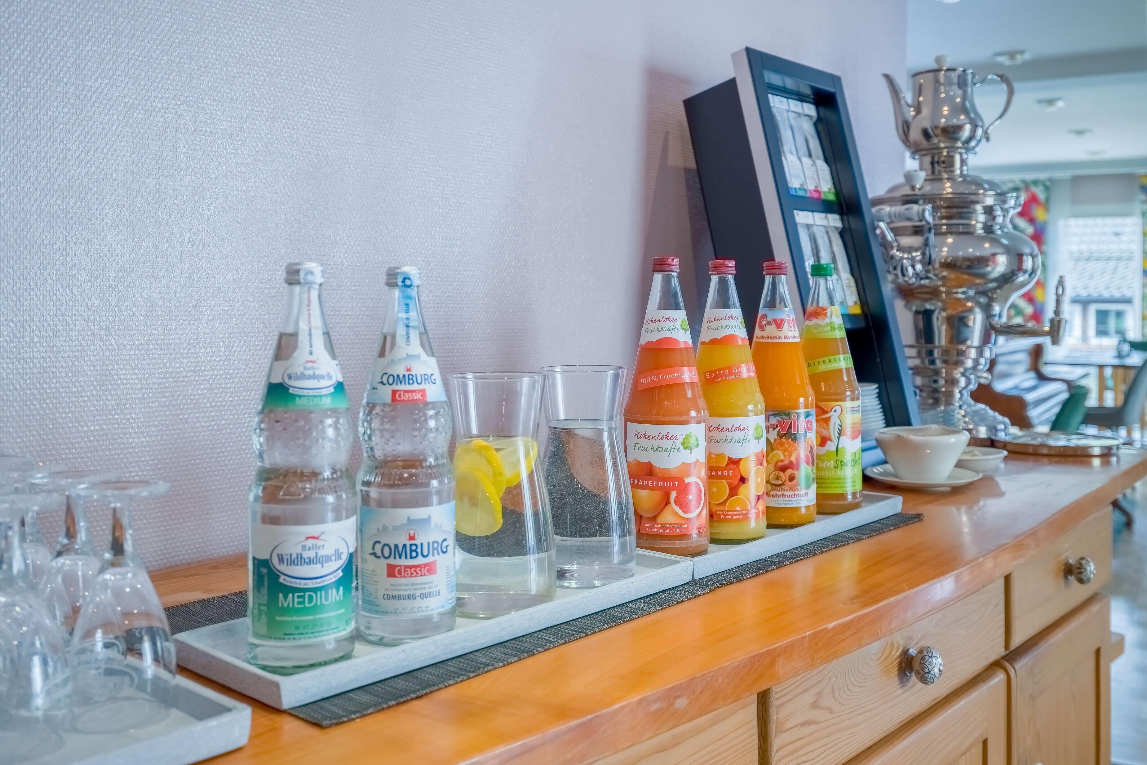 Un espace bar avec différentes bouteilles d’eau et de jus, des verres et un pichet décoratif en argent.