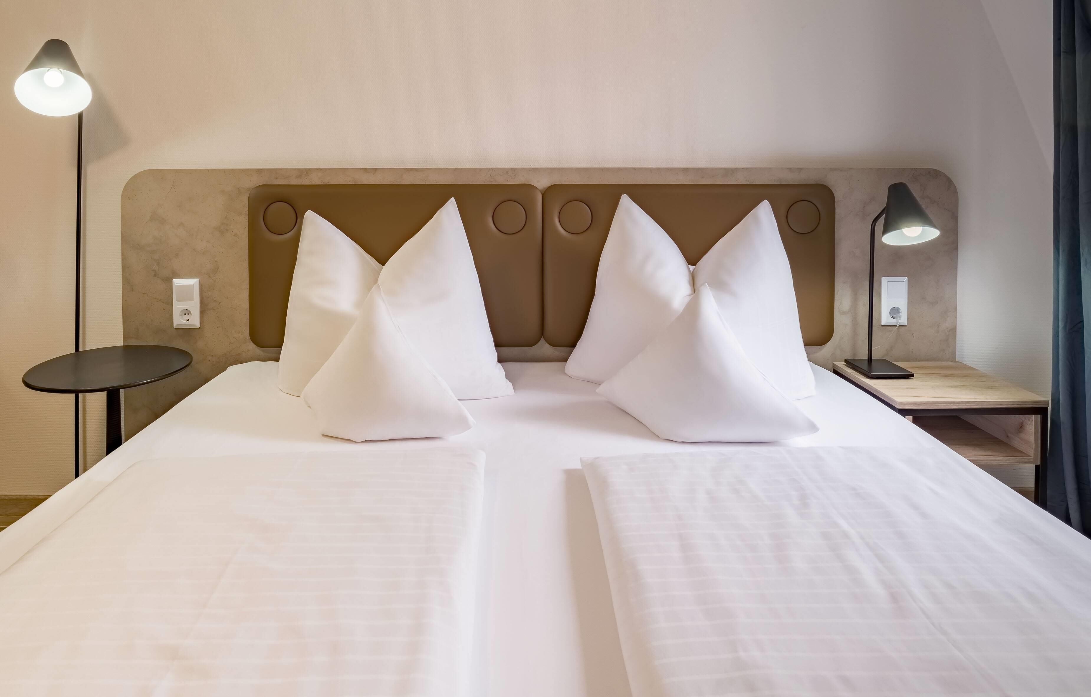 A neatly made bed with white linens and pillows, a bedside table with a lamp, and a wall-mounted light fixture.
