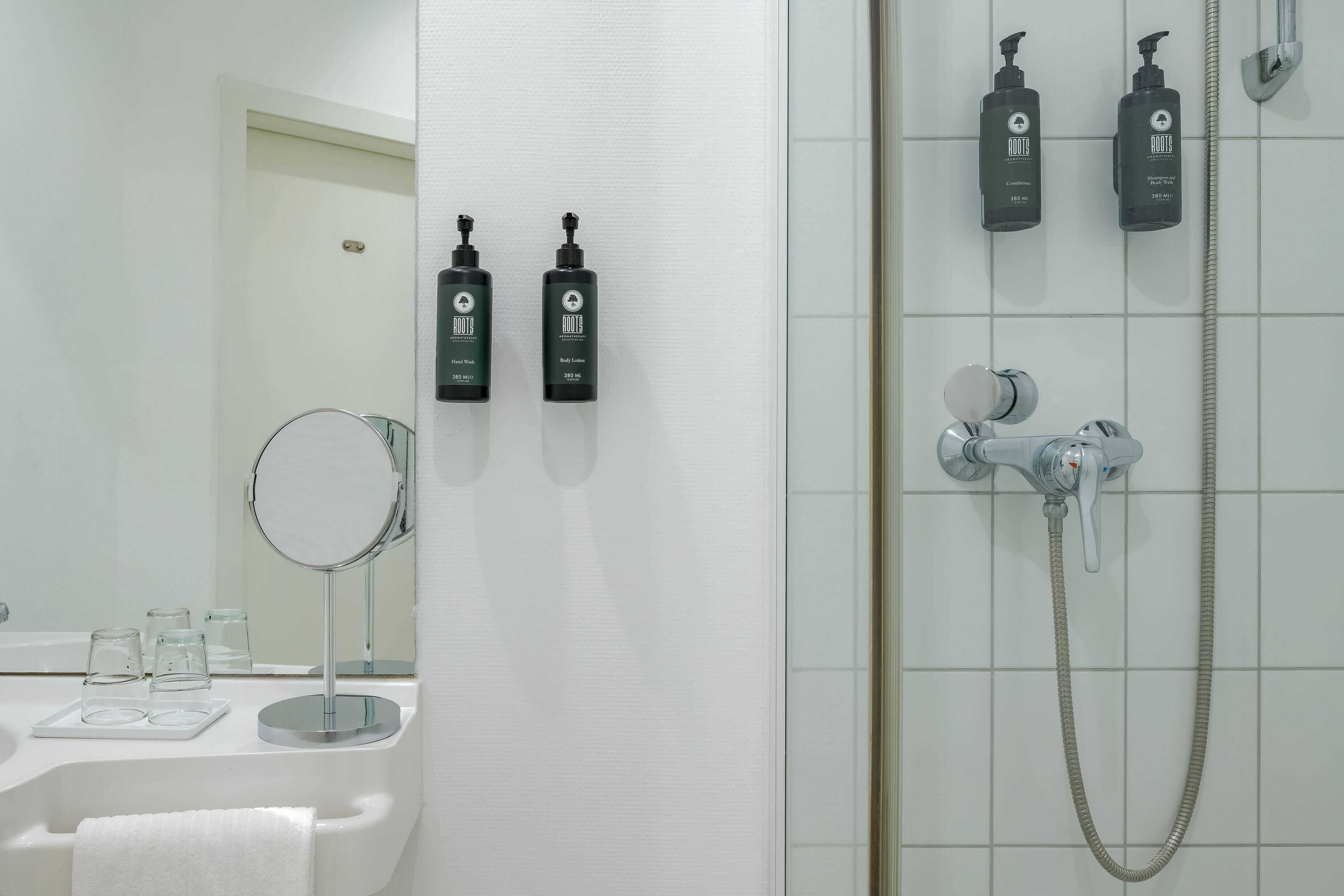 A bathroom with a white sink, a round mirror, two bottles of Roots hand wash and body lotion, and a shower with a handheld showerhead and hose.