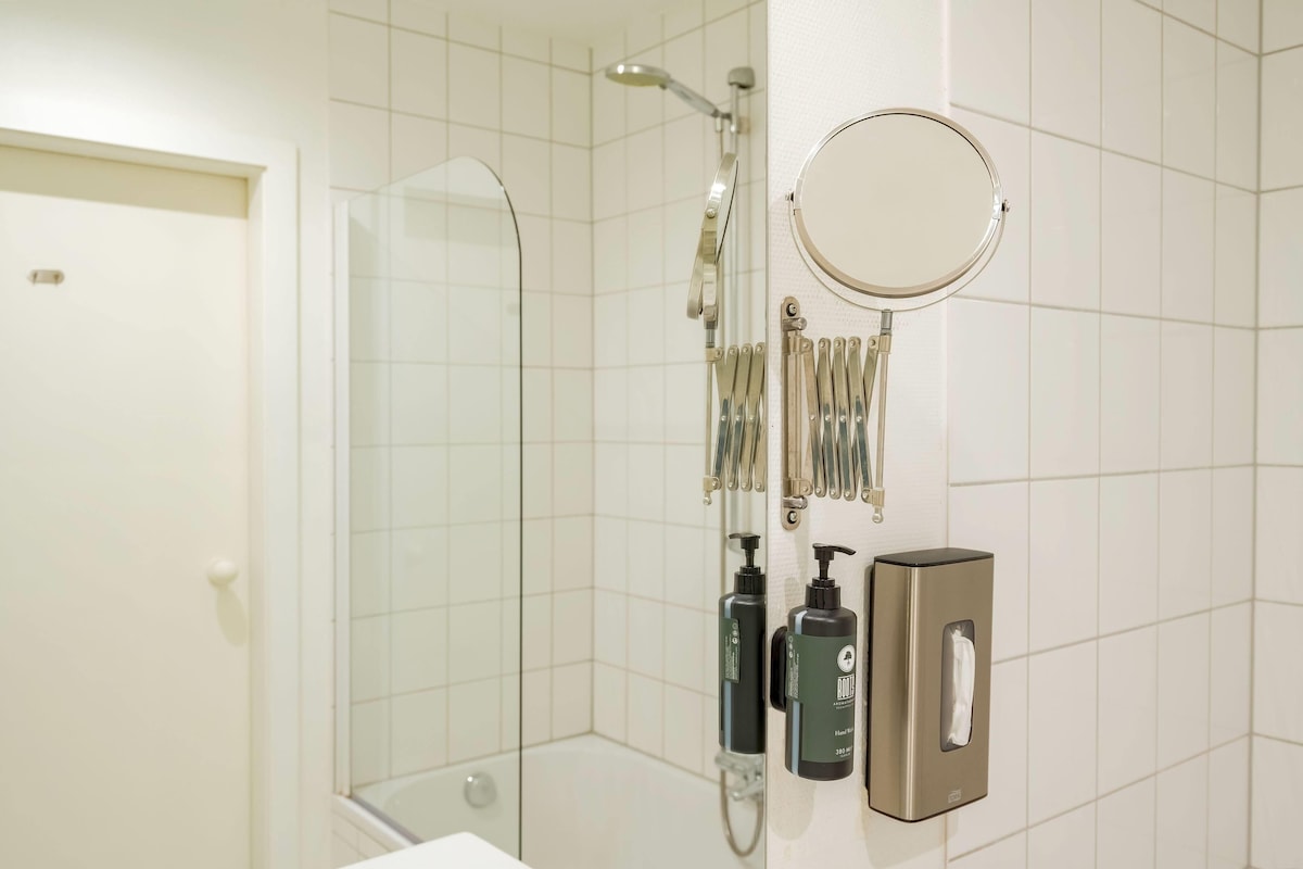 A modern bathroom with a glass shower enclosure, a handheld showerhead, a wall-mounted mirror, and toiletries.