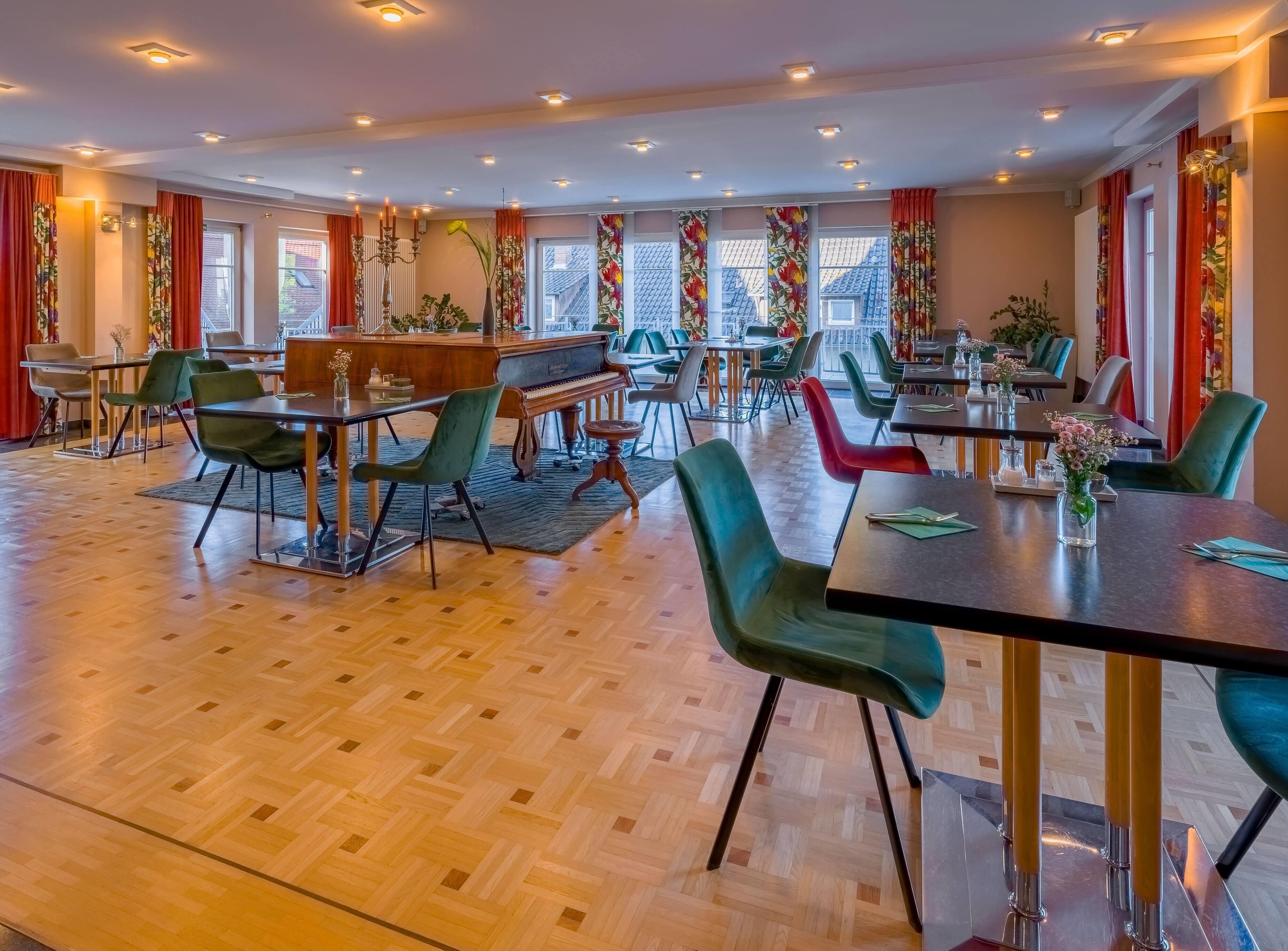A spacious dining area with multiple tables and chairs, a piano, and large windows with red curtains.