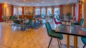 A spacious dining area with multiple tables and chairs, a piano, and large windows with red curtains.