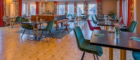 A spacious dining area with multiple tables and chairs, a piano, and large windows with red curtains.
