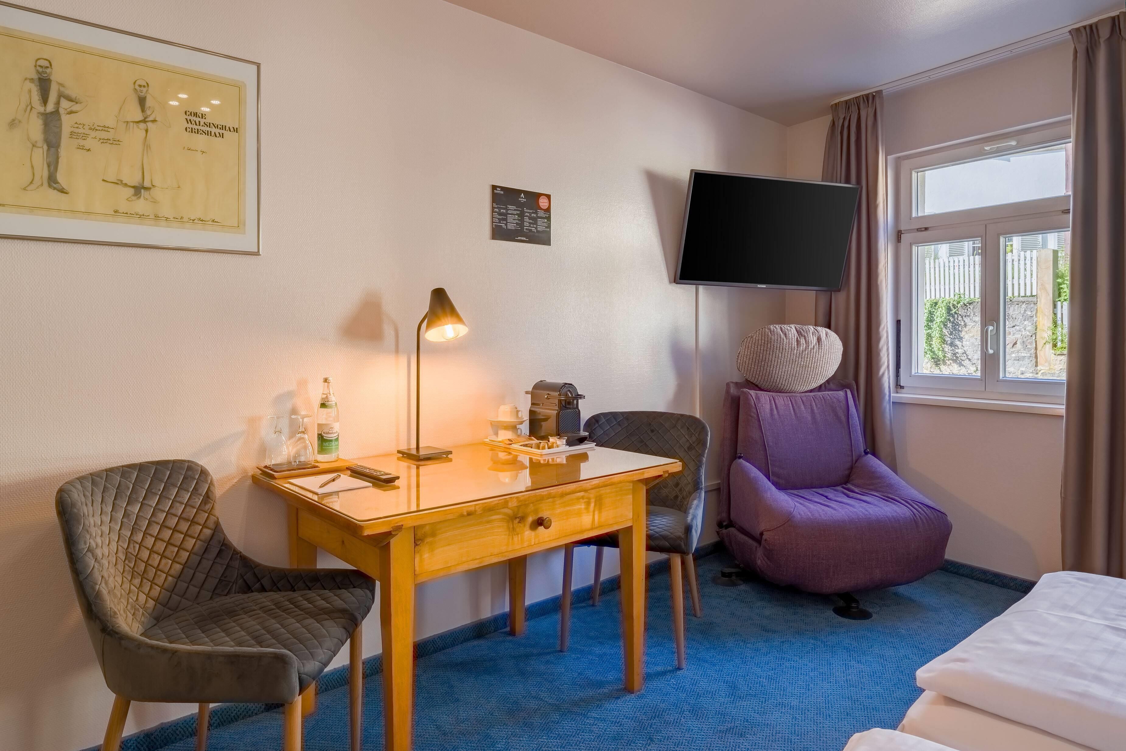 Habitación de hotel con cama, escritorio con lámpara, televisión y un sillón morado.