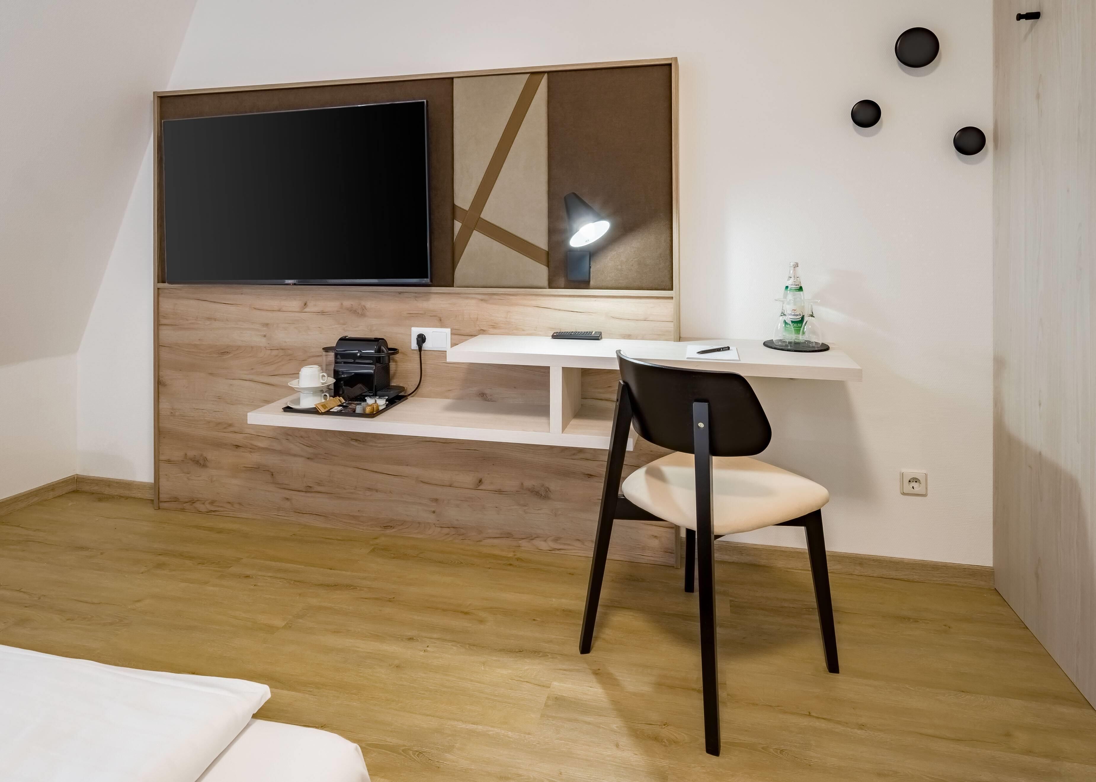 Une chambre moderne avec un bureau en bois, une chaise, un téléviseur fixé sur un panneau en bois, une lampe de bureau et une petite étagère sur laquelle se trouvent une bouteille et une télécommande.