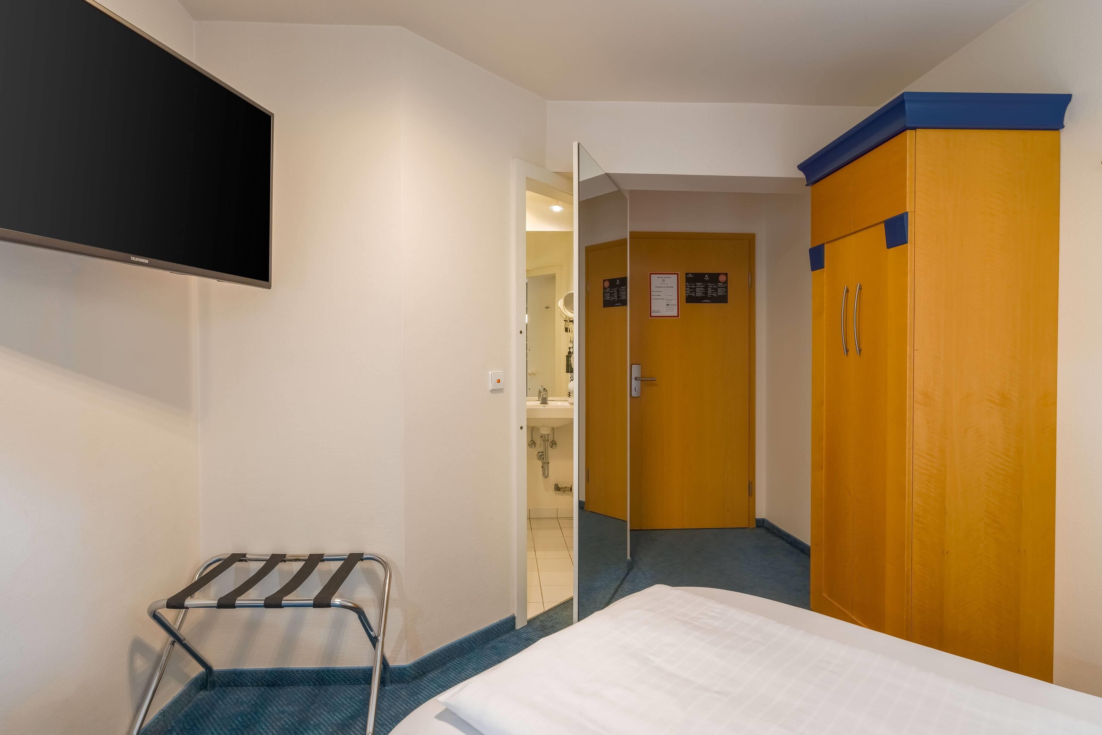 Habitación de hotel con cama, televisor montado en la pared, armario y baño visible a través de una puerta abierta.
