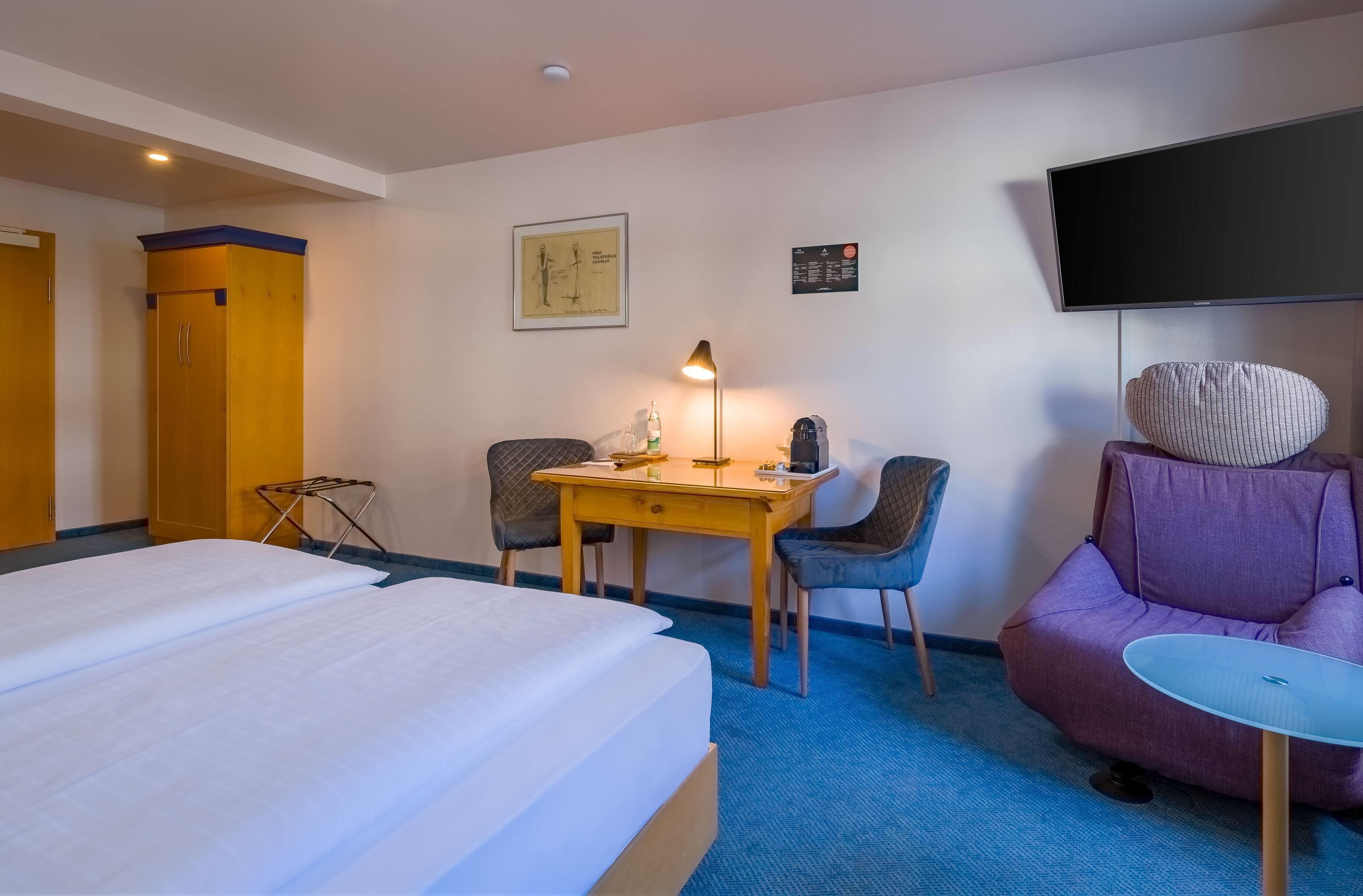 Una habitación de hotel con una cama, un escritorio con lámpara, dos sillas, un sillón morado y un televisor en la pared.