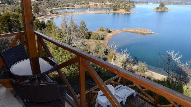 Um deck de madeira com mesa e cadeiras, oferecendo vista para um lago com ilhas e montanhas ao longe.