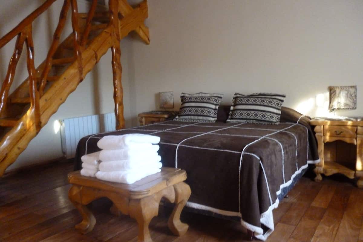 Un dormitorio con una escalera de madera, una cama con cubrecama marrón oscuro y almohadas blancas, una mesita de madera con toallas dobladas y una mesita de noche con una lámpara.