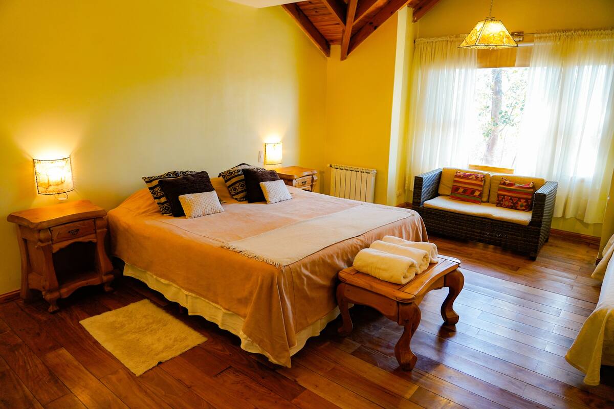 Un dormitorio con una cama grande, mesitas de noche, un banco y una ventana con cortinas.
