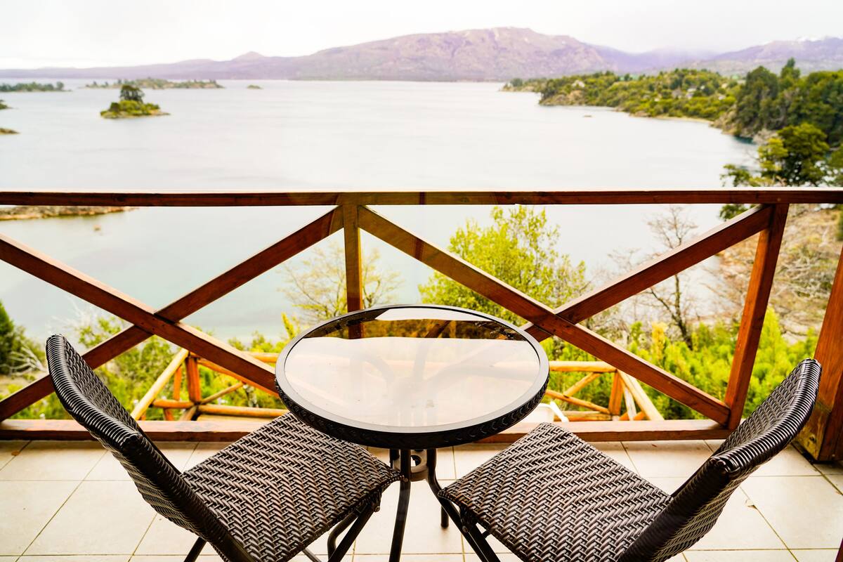 Un balcón con una mesa y dos sillas, con vista a un lago con islas y montañas a lo lejos.