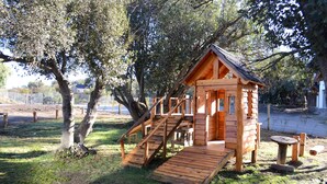 Uma casinha de madeira com rampa e escadas, rodeada por árvores e uma área cercada.