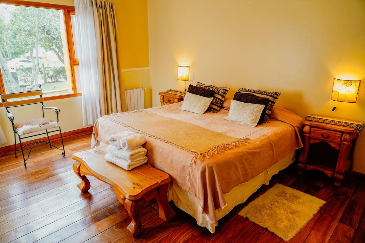 Un dormitorio ordenado con una cama grande, mesitas de noche, un banco y vistas al exterior.