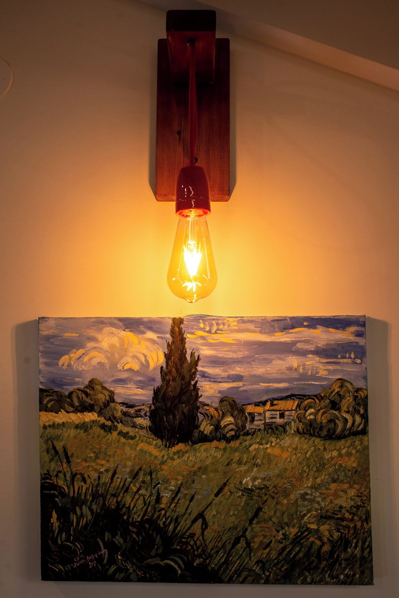 Una lámpara de pared con base de madera y bombilla de vidrio, iluminando un cuadro de paisaje abajo.