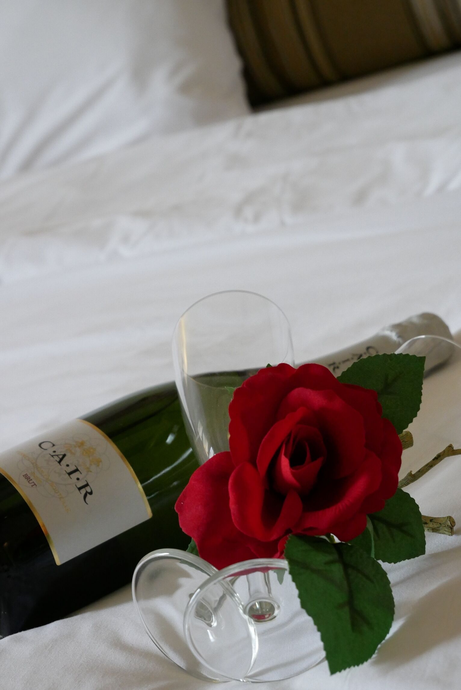 A bottle of wine, a glass, and a red rose on a bed.
