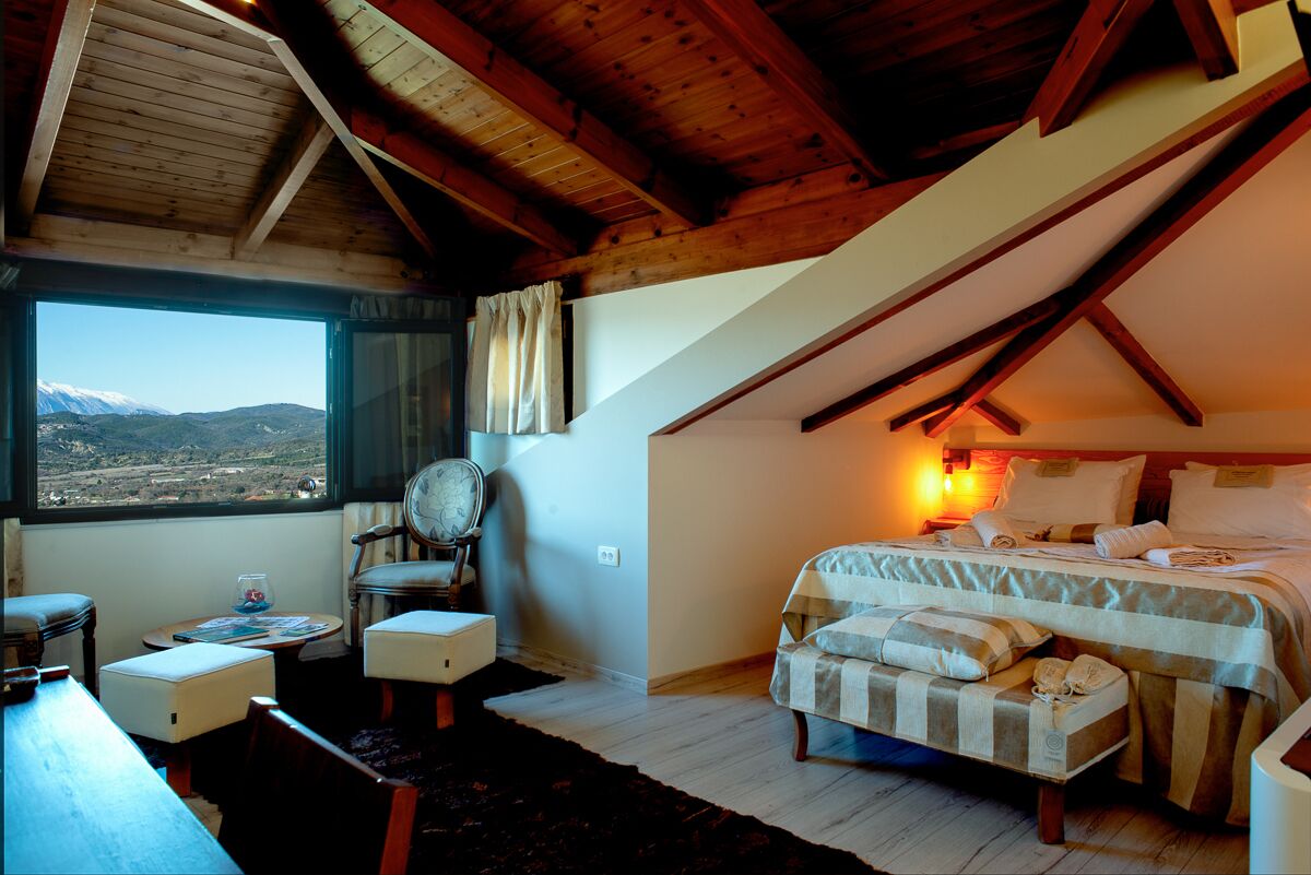 A bedroom with a bed, a chair, a table, and a window with a view of the mountains.