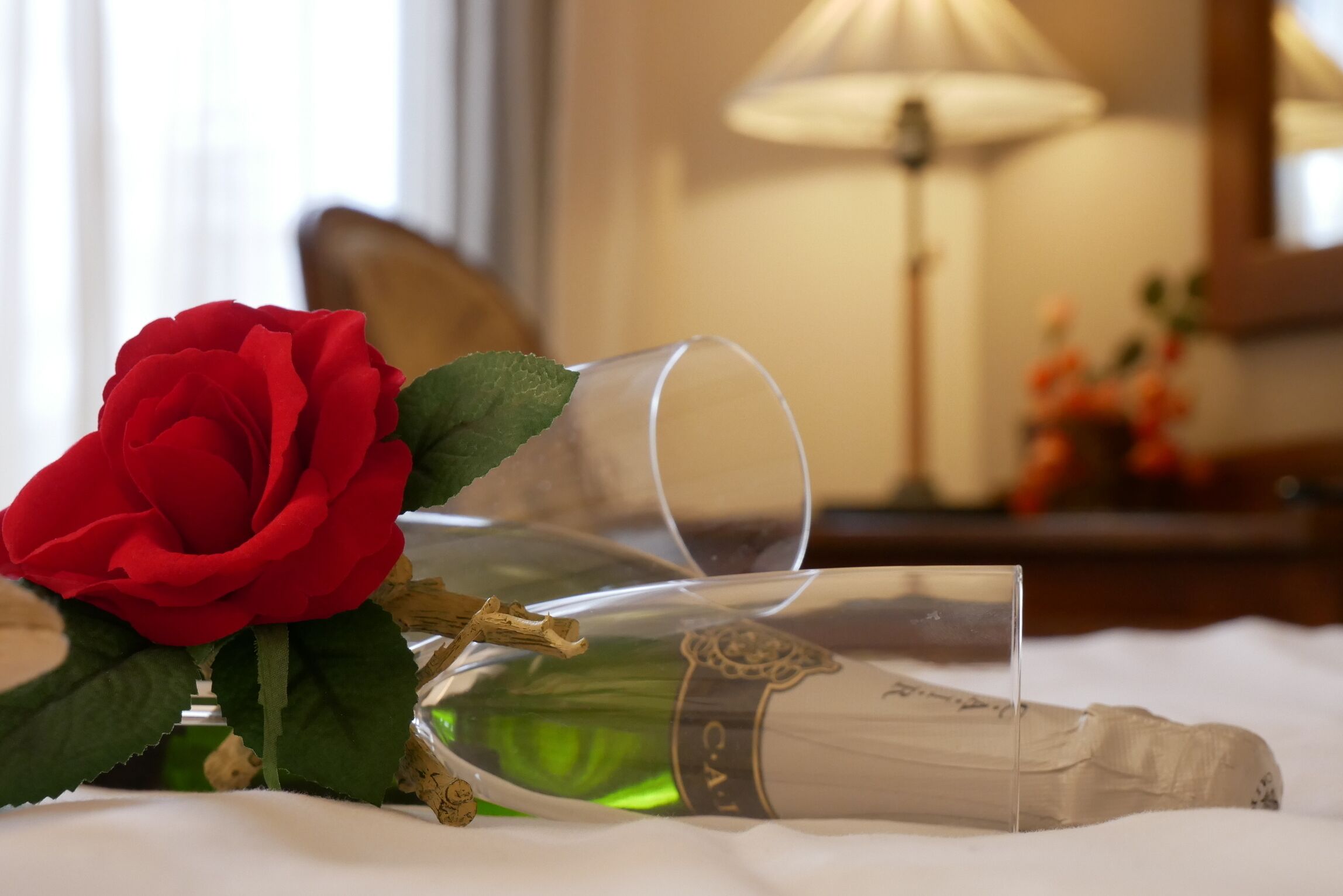 A champagne bottle and a red rose on a bed.