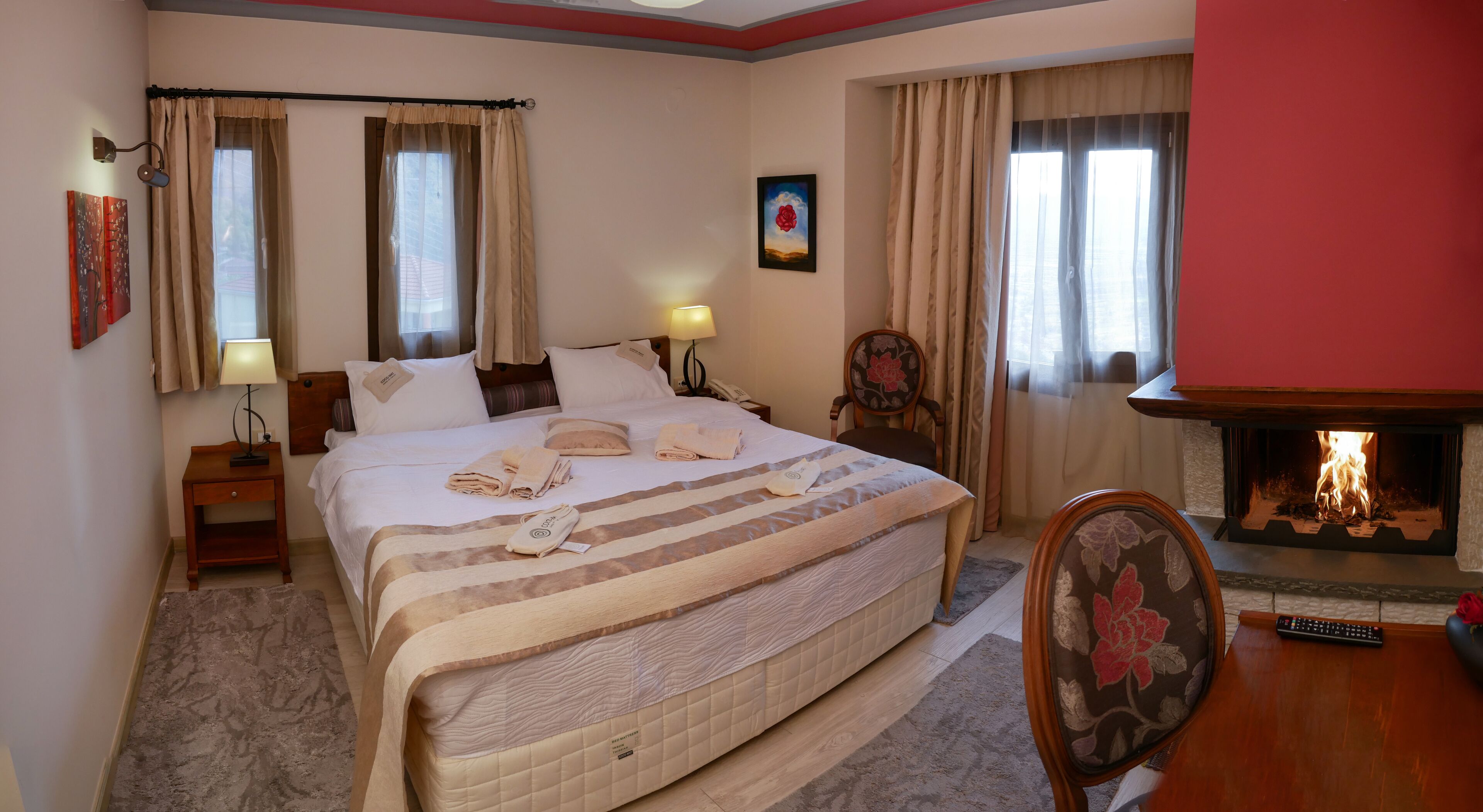 A hotel room with a large bed, a bedside table with a lamp, a chair, a fireplace, and a painting on the wall.