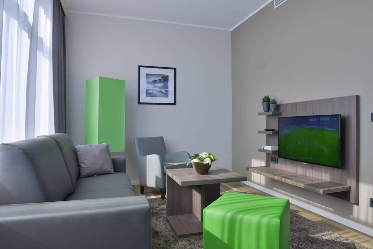 Un salon moderne avec un canapé gris, une table basse en bois, un pouf vert, un téléviseur fixé au mur et un tableau encadré accroché au mur.