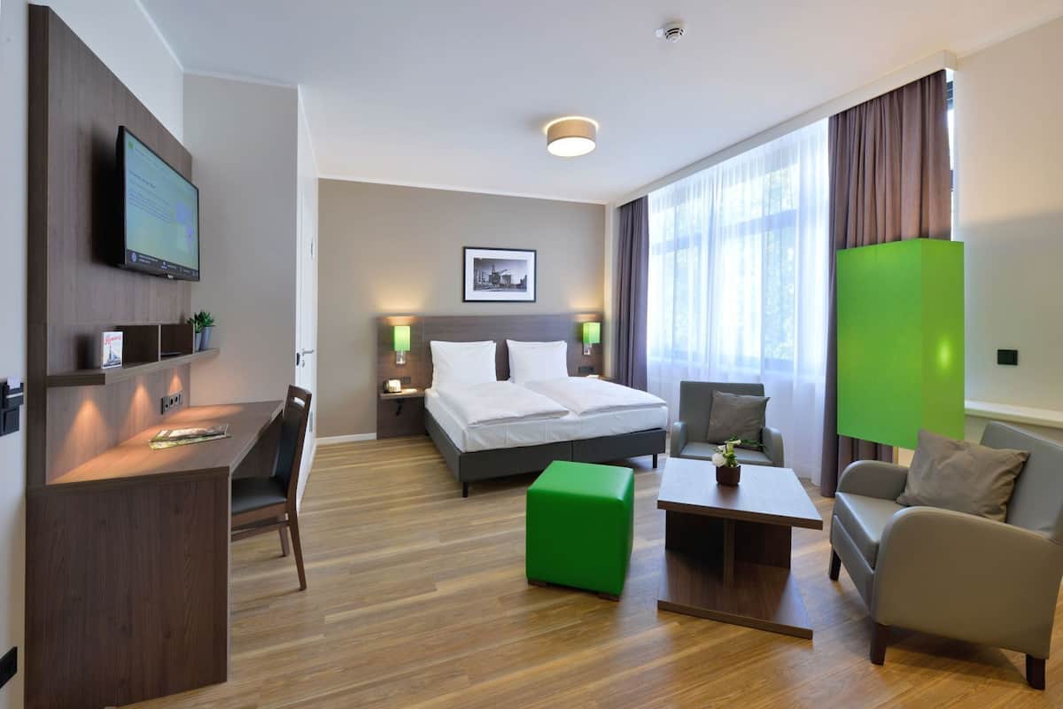 Une chambre d’hôtel comprenant un lit, un bureau, une chaise, un pouf vert et un canapé.