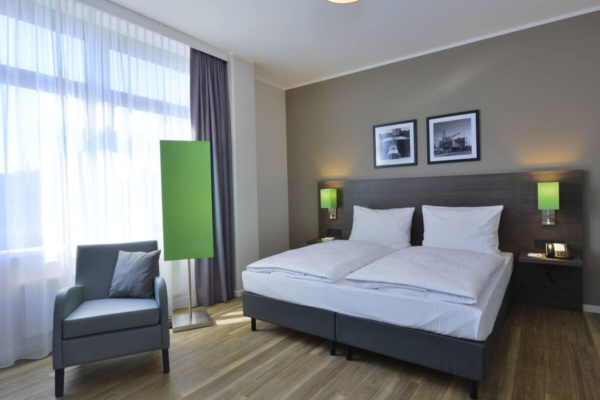Une chambre d’hôtel avec un lit, une chaise, une table de chevet, une lampe et une fenêtre avec des rideaux.
