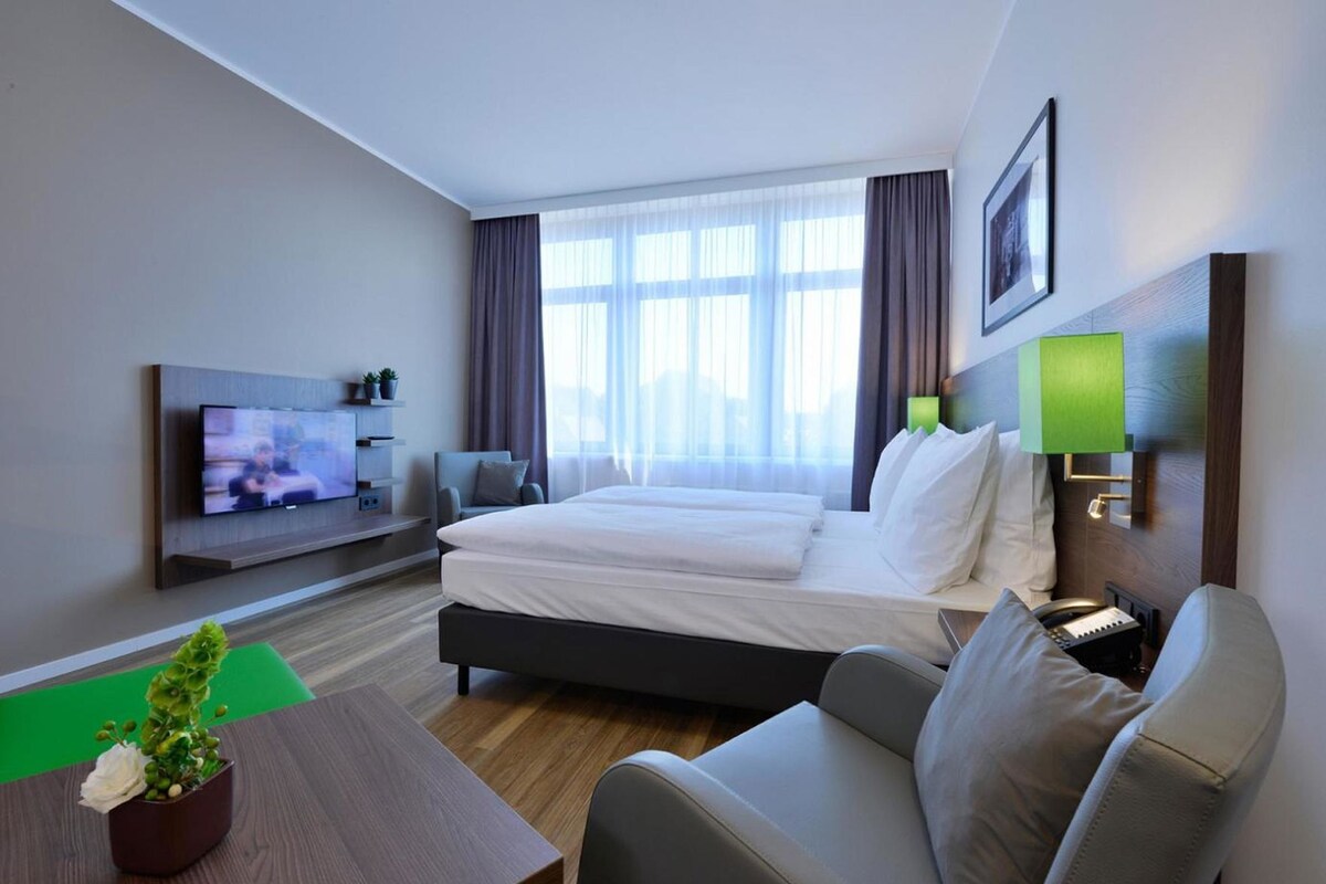 Hotelkamer met een bed, een televisie, een bank, een tafel met een plant en een raam met gordijnen.